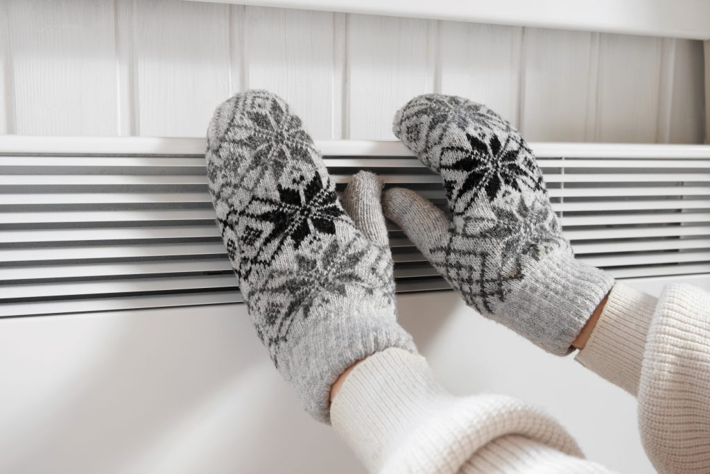 Hands in winter gloves warming up over electric heater. Frozen woman wearing a knitted mittens freezing for winter cold. Discomfort spending time at home. Girl warming hands on modern radiator.